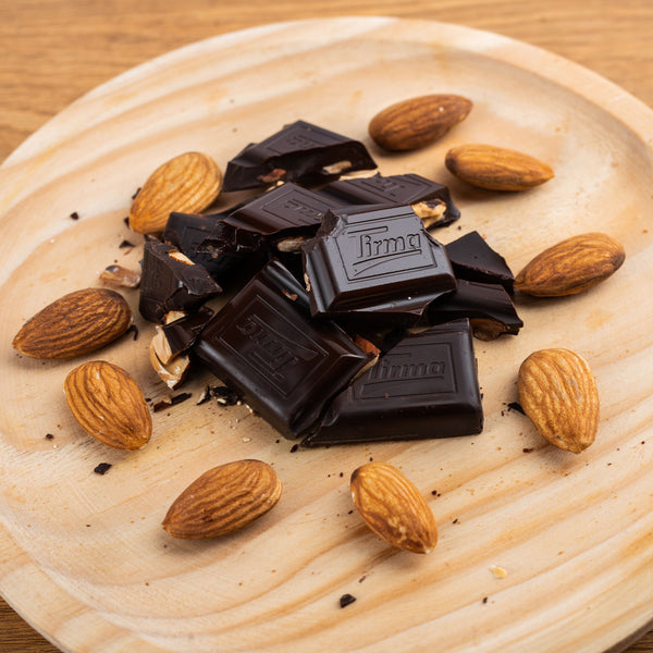 Tirma 70% dark chocolate bar with whole almonds, 125g. Gluten Free placed in a wooden plate with almonds around it. Spanish chocolate with almonds made in Spain.