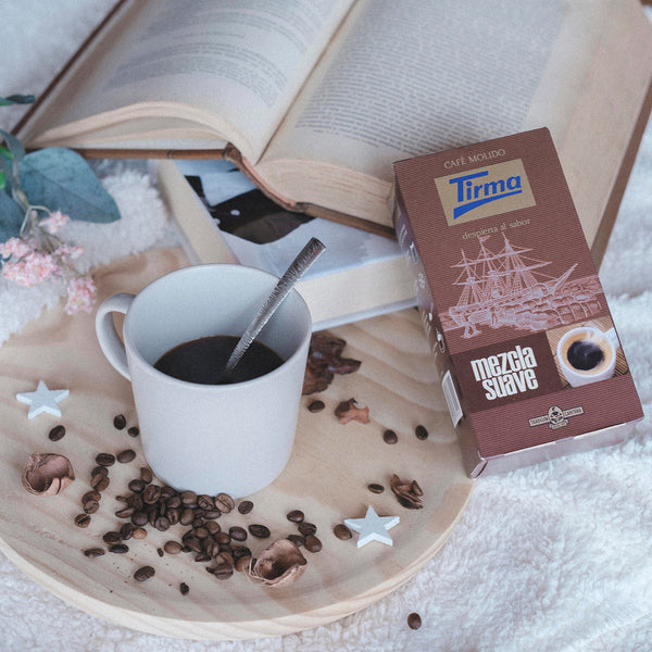 Tirma Soft Ground Blend Coffee placed next to a book, scattered coffee beans and a cup of coffee with spoon. Spanish Coffee made in Spain.