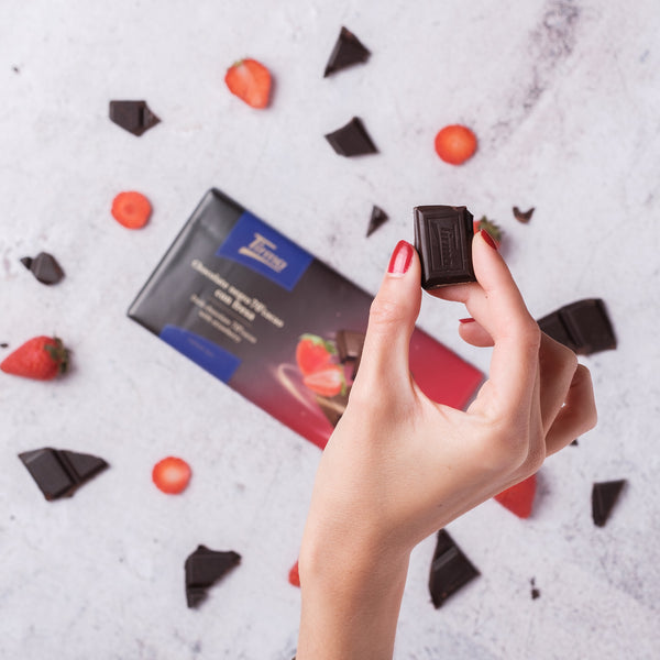 Tirma 70% Dark Chocolate with Strawberry Granules held by a hand and scattered strawberries. Spanish strawberry chocolate made in Spain.