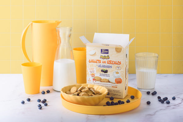 Tirma Cereal and Blueberry Mini Biscuit Semicovered with White Chocolate 160 g filled in the yellow bowl in a yellow serving platter placed next to glass and milk with yellow pitcher and scattered blueberries. Spanish biscuits made in Spain.