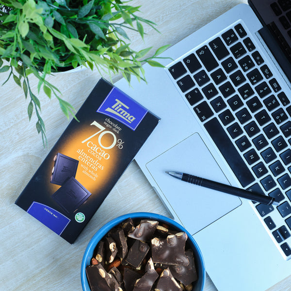 Tirma 70% dark chocolate bar with whole almonds, 125g. Gluten Free Chocolate placed next to the laptop and a ballpen. Spanish chocolate with almonds made in Spain.