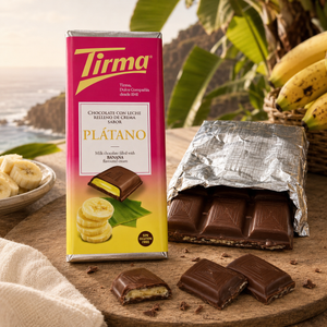 A package of Tirma banana chocolate bar sits on a wooden table with a partially unwrapped chocolate bar in foil and several chocolate pieces in front. Bananas and a bowl are visible in the softly blurred background.