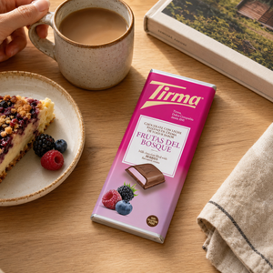 A Tirma Frutas del Bosque chocolate bar in a pink wrapper, styled on a wooden table next to a cup of coffee, a slice of berry pie on a plate, and a folded cloth.