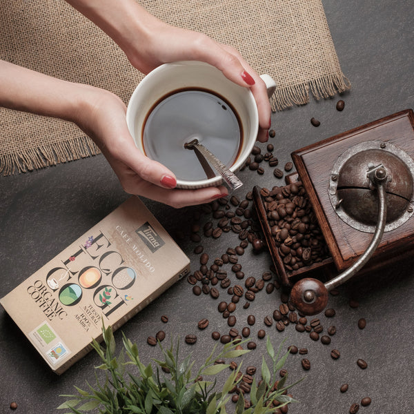 Tirma Organic Blend Coffee 250 g placed next with a hands holding the cup of coffee with a spoon and scaterred coffee beans. Spanish coffee made in Spain.